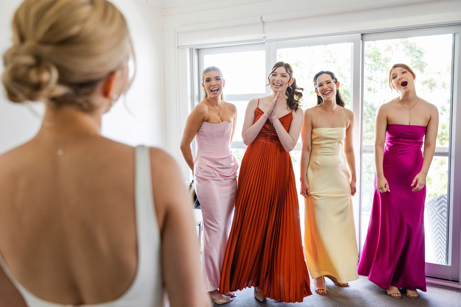 Bridesmaids in colorful satin dresses react with joy and excitement while looking at someone in a bright room.