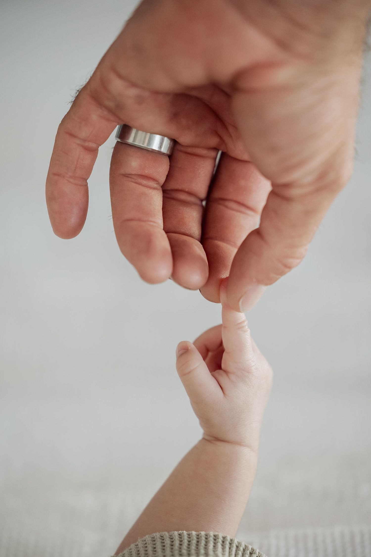 A small baby's hand reaches up to touch an adult hand wearing a silver ring against a light background.
