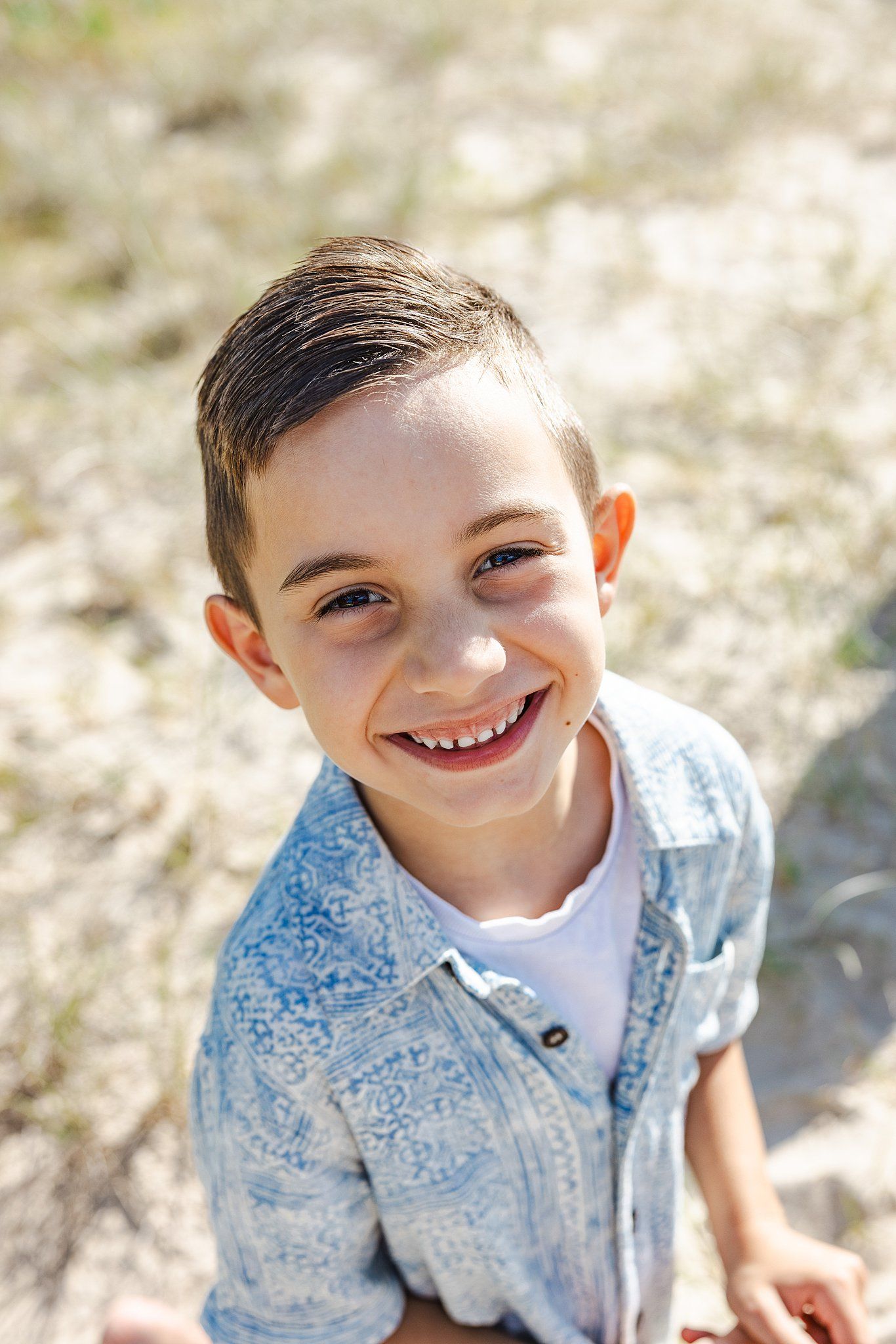 Cheerful young child wearing a light blue patterned button-down shirt smiles brightly in outdoor sunlit setting.