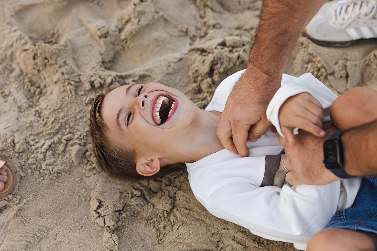 Playful moment captured at the beach as someone lies in the sand being tickled and laughing joyfully.
