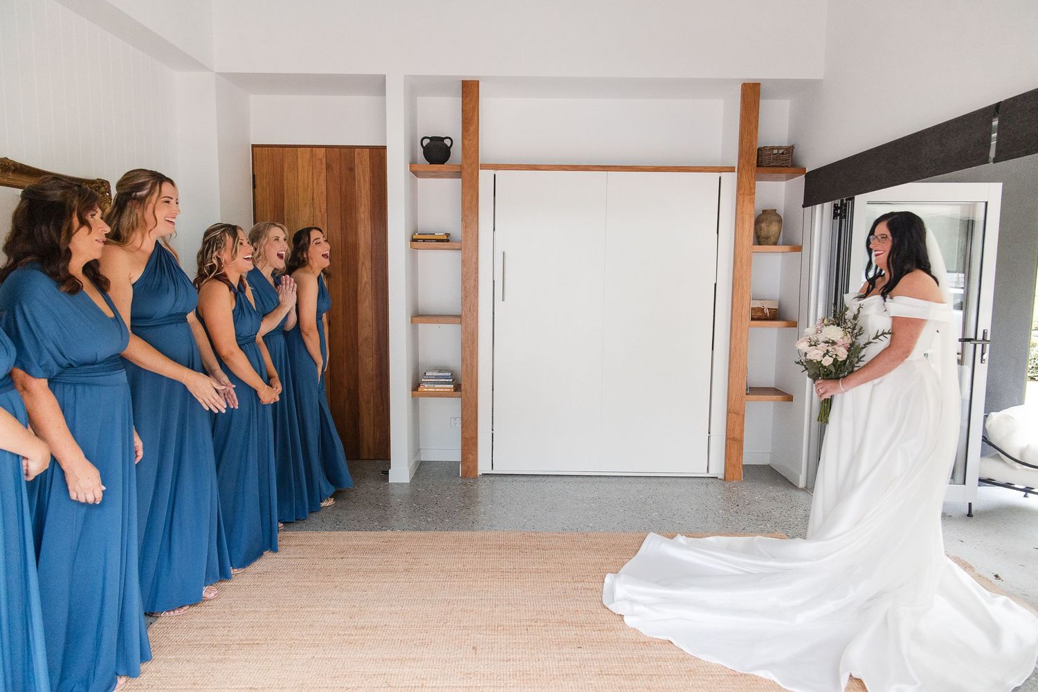 Bride in white gown reveals herself to bridesmaids in blue dresses during wedding morning first look.