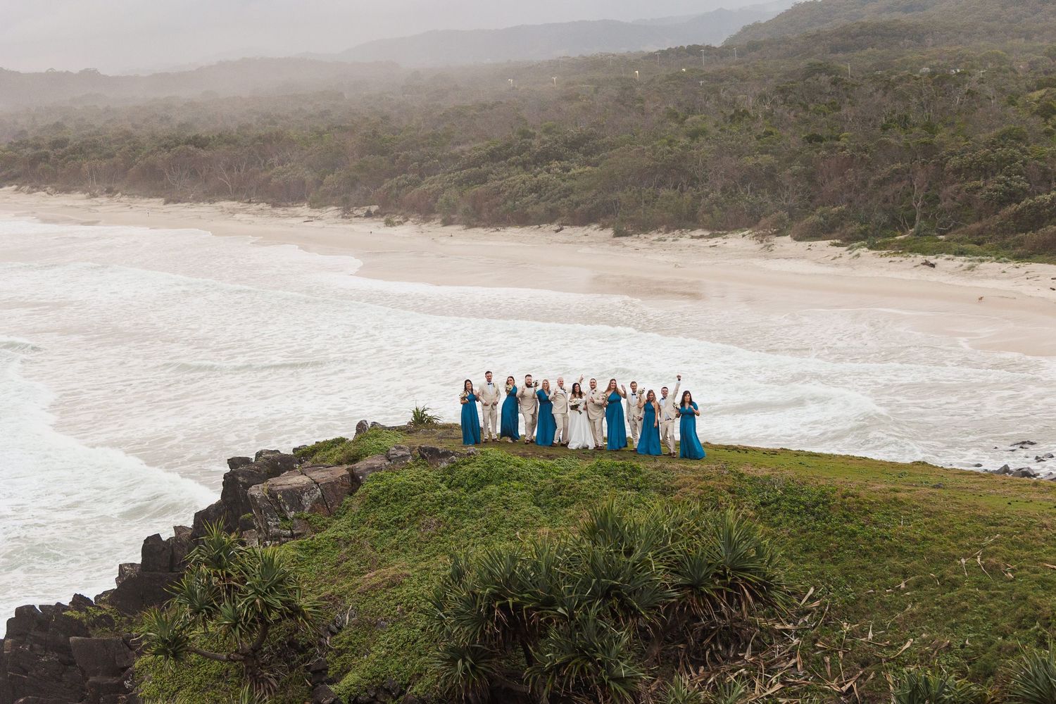 Bridal party in teal gowns standing on coastal cliff overlooking a misty beach with rolling waves and lush greenery.