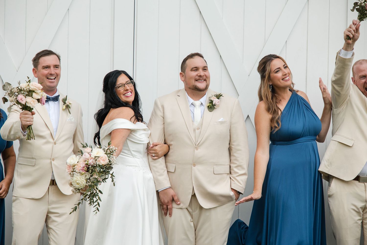 Wedding party in beige suits and blue dress celebrating joyfully outside a white barn venue.