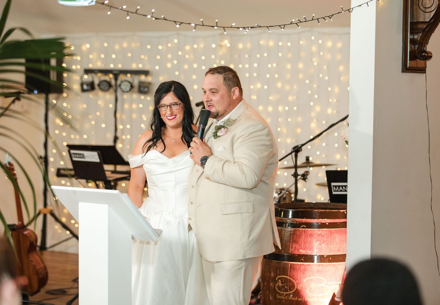 Bride and groom smiling together at wedding ceremony with fairy light backdrop and live band in elegant venue.