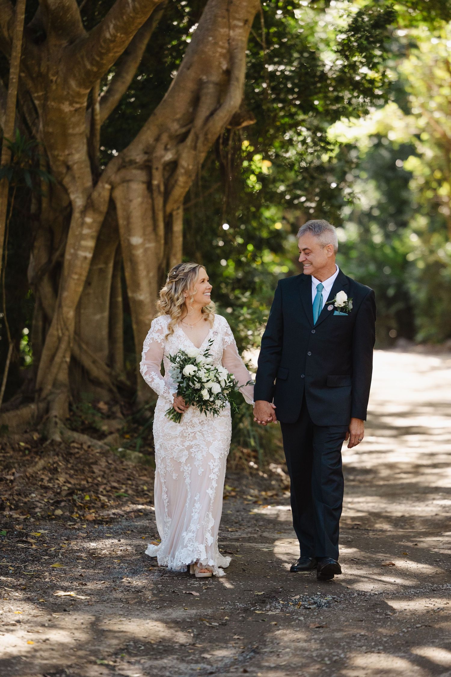 A bride in a lace wedding dress walks along a shaded garden path with an older gentleman in a navy suit.