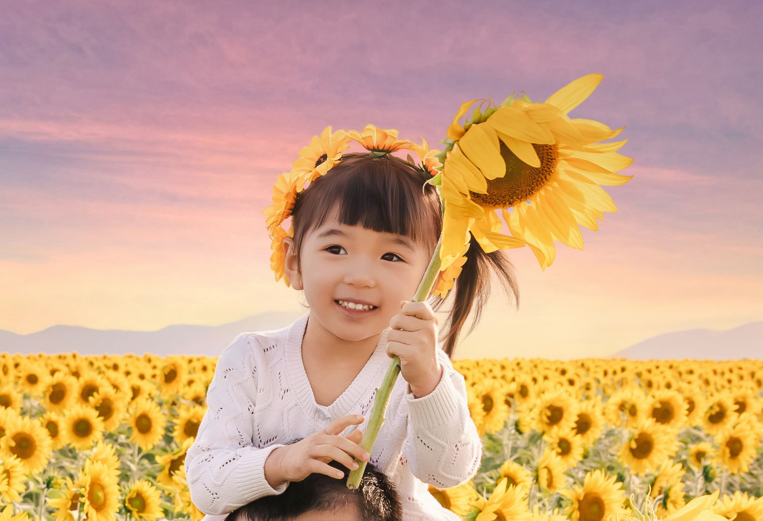 Smiling young girl holding a sunflower in a vibrant sunflower field wearing a floral crown at sunset.