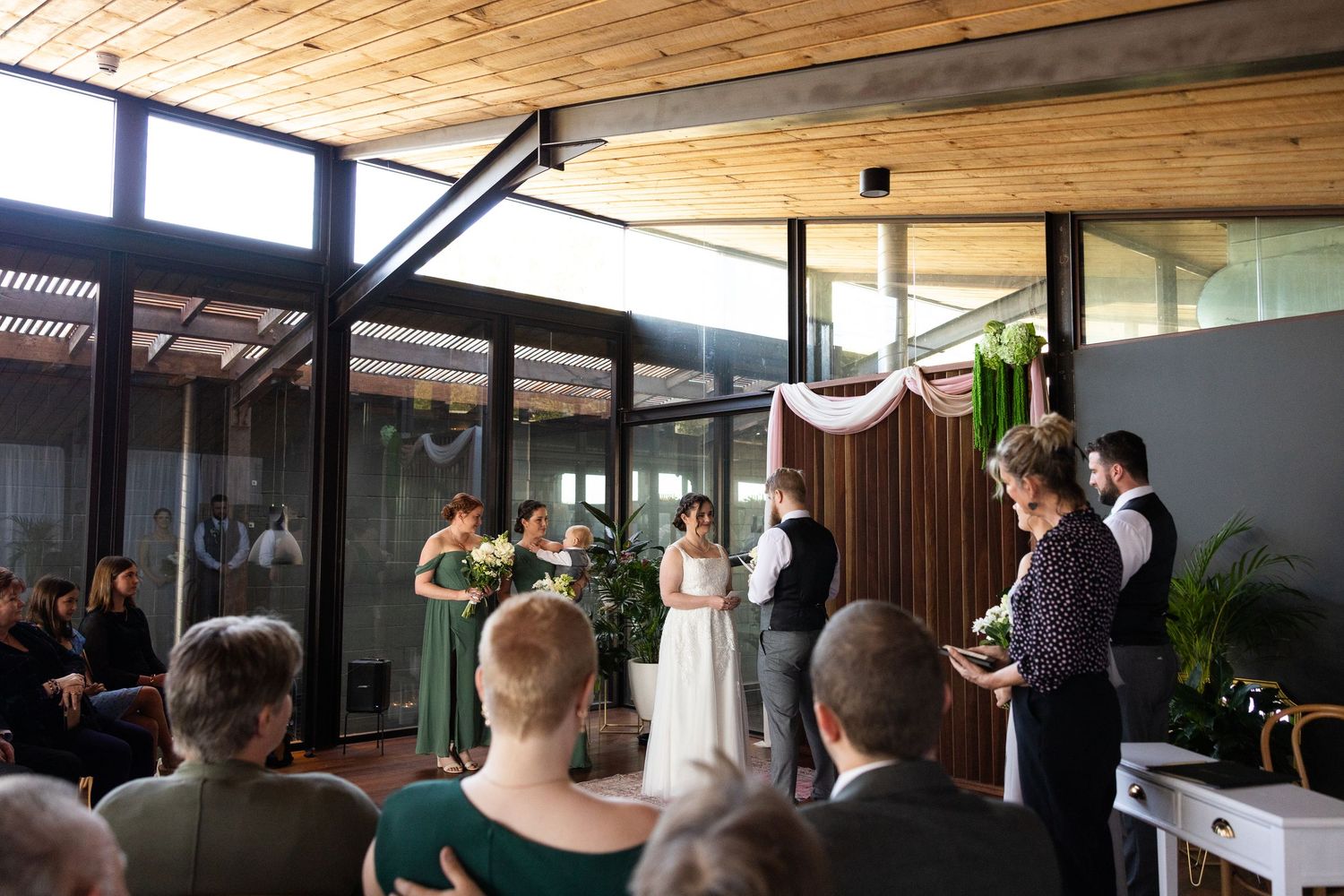 Intimate indoor wedding ceremony with couple exchanging vows before officiant, guests seated, modern venue with wood ceiling.
