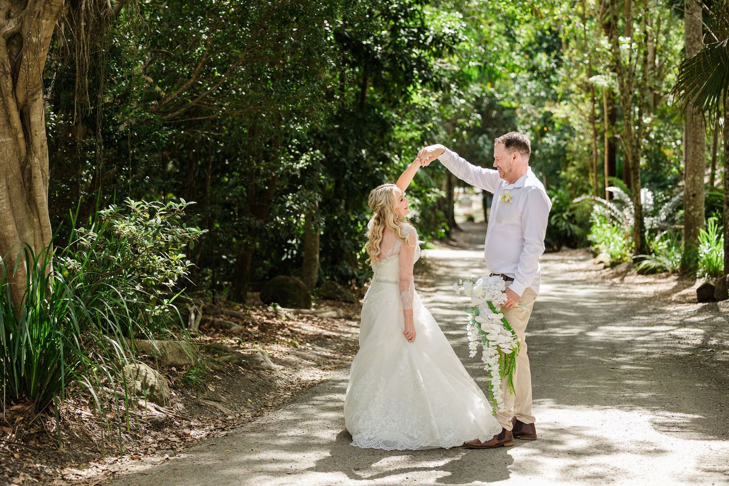 Bride and groom dancing on a tree-lined path outdoors during a romantic wedding celebration.