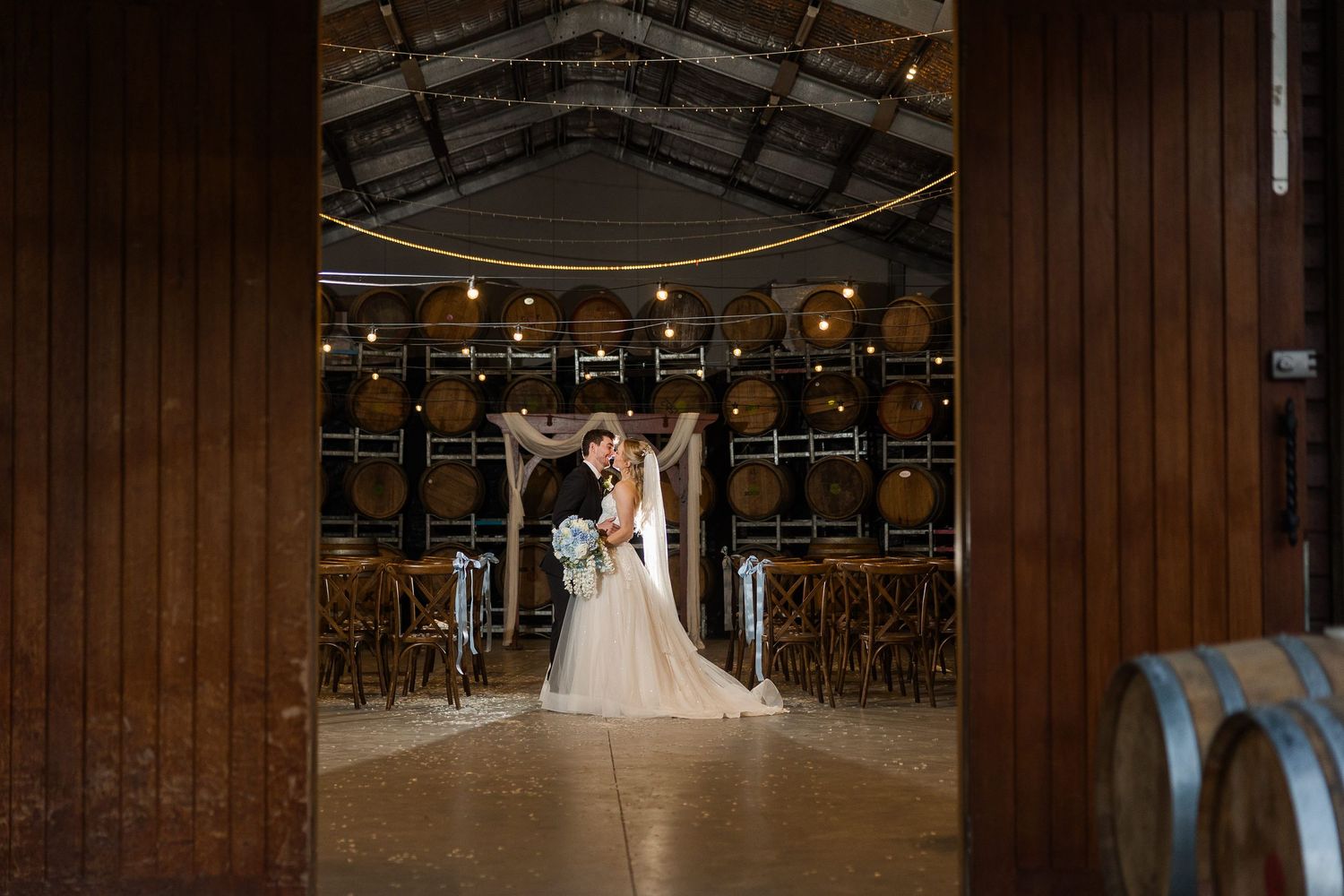 Bride and groom share a romantic moment in a rustic winery barrel room decorated for an elegant wedding ceremony.
