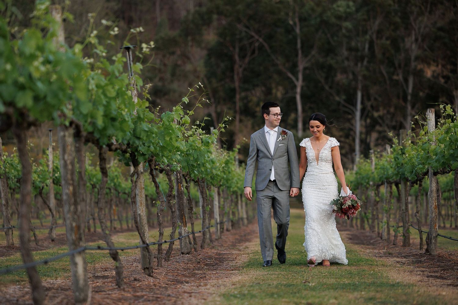 Bride and groom walking hand in hand through lush green vineyard rows on their wedding day.