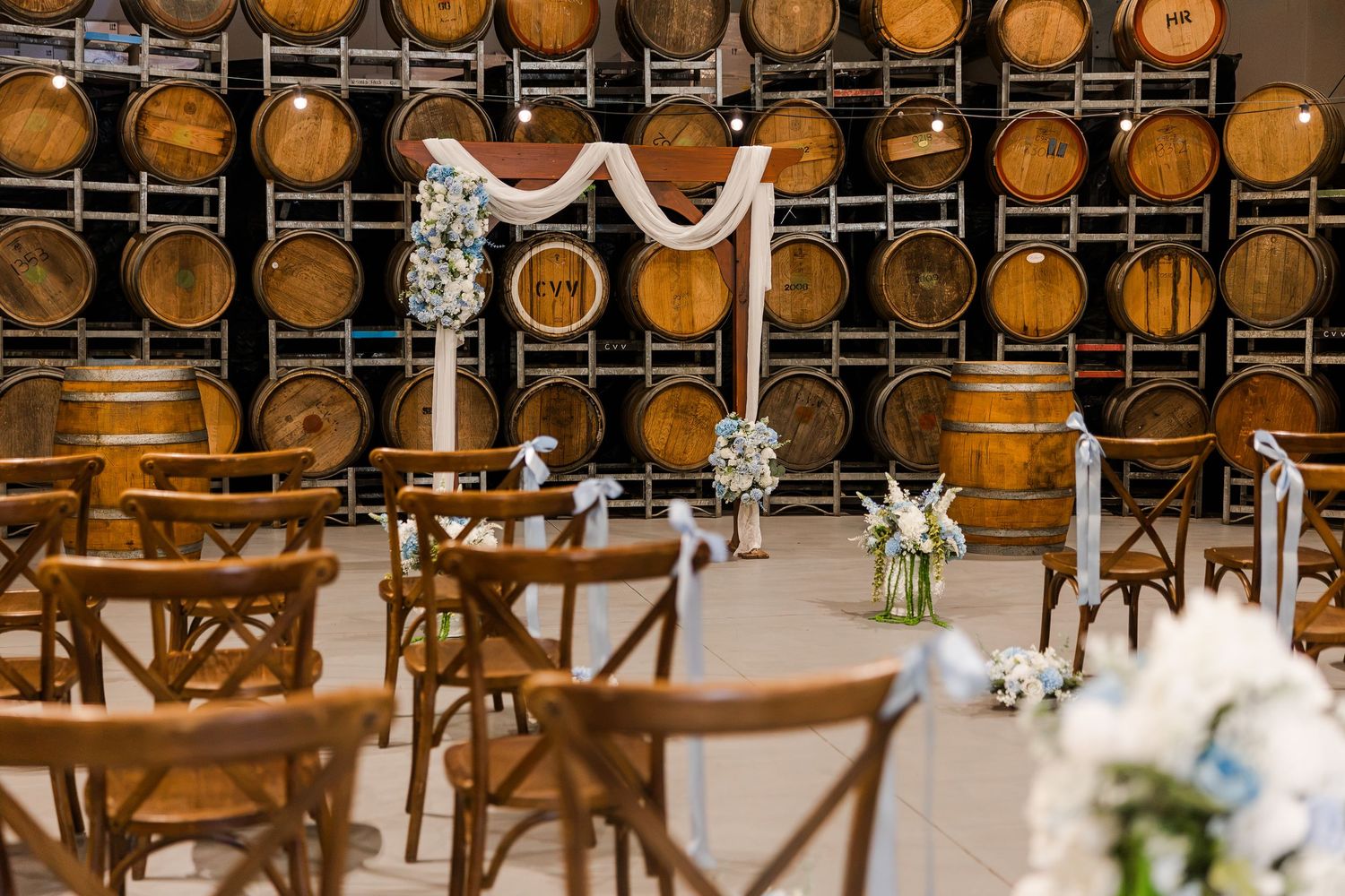 Elegant winery wedding ceremony setup with wooden barrel wall backdrop, floral arch, cross-back chairs, and white flower arrangements.