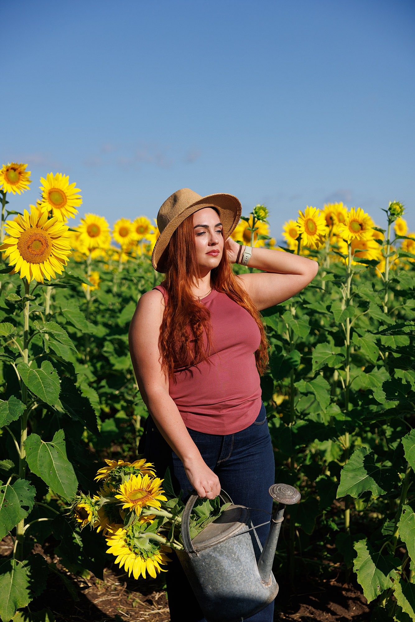 Standing in a bright sunflower field on a sunny day wearing a straw hat and burgundy top with a metal watering can.