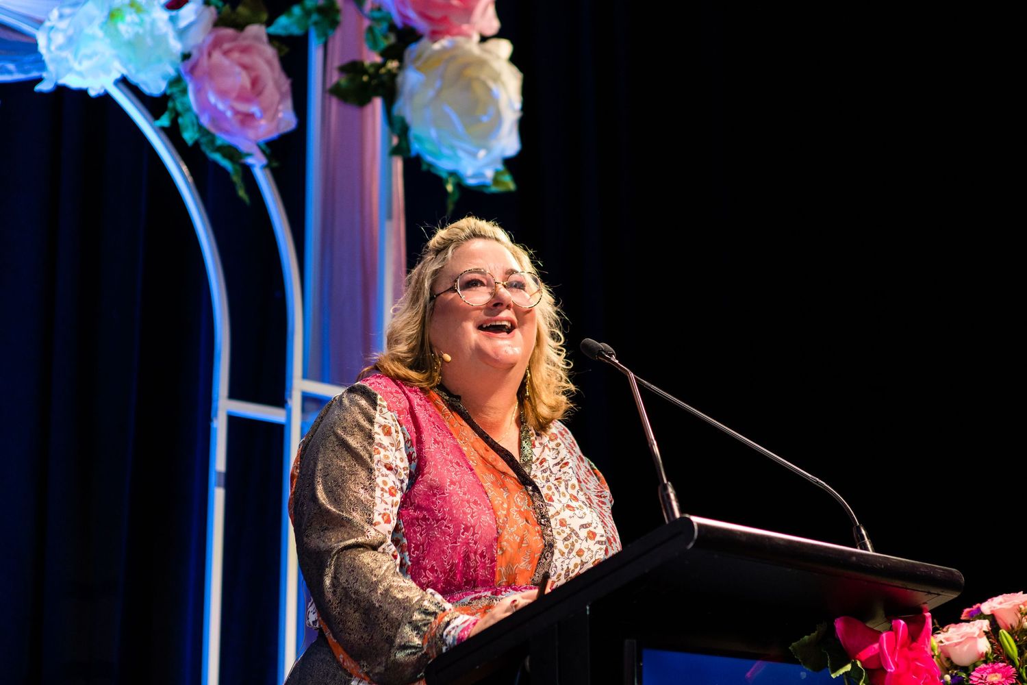 A speaker stands at a podium giving a presentation at an event decorated with pink and blue floral arrangements.