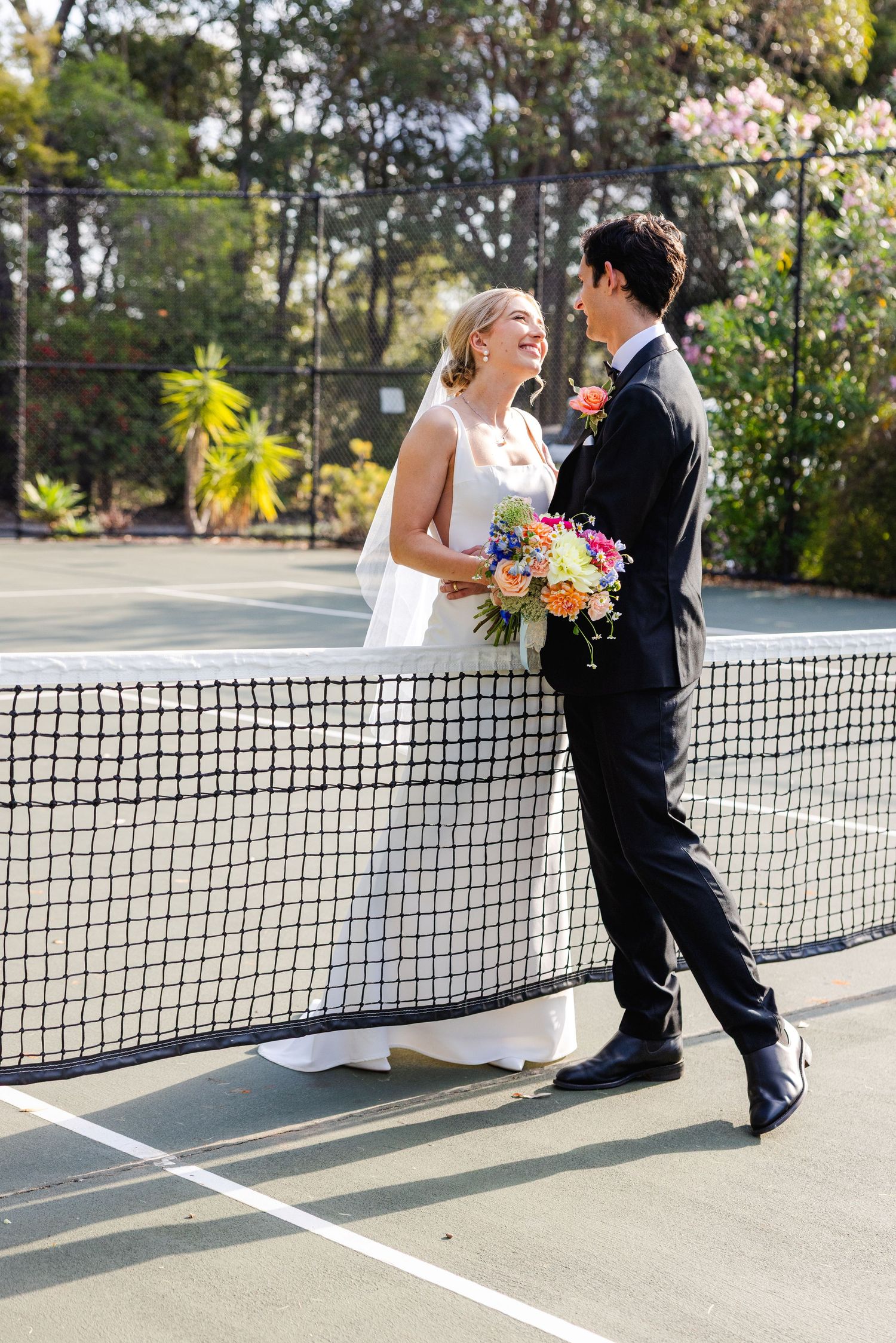 A couple in wedding attire shares a playful moment on a tennis court with a bouquet and net between them.