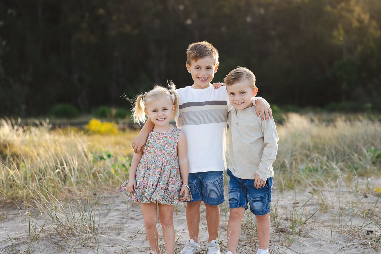 Three young children smiling and posing together outdoors on a sandy path surrounded by golden grass.
