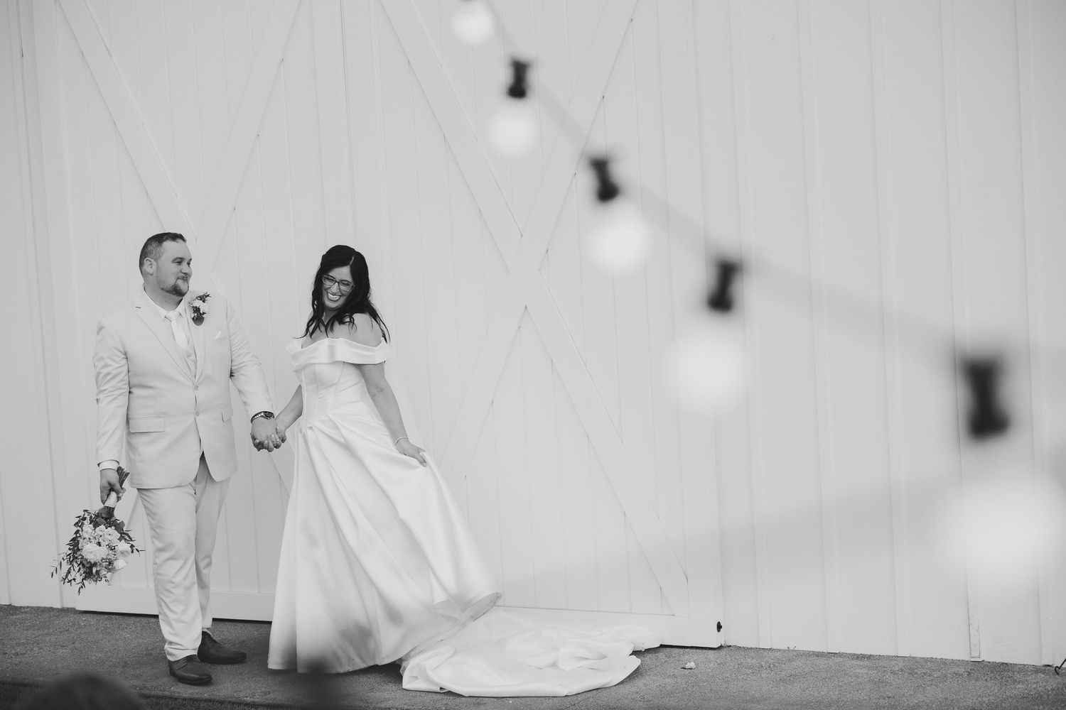 Black and white photo of a bride and groom holding hands at a barn wedding venue with string lights.