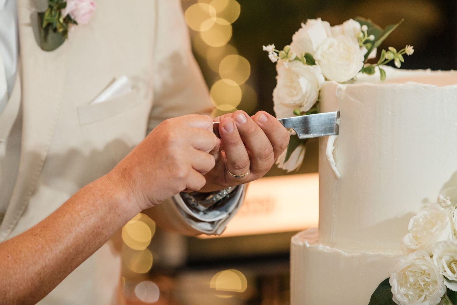 Couple cutting a white wedding cake decorated with fresh roses and greenery at a reception.