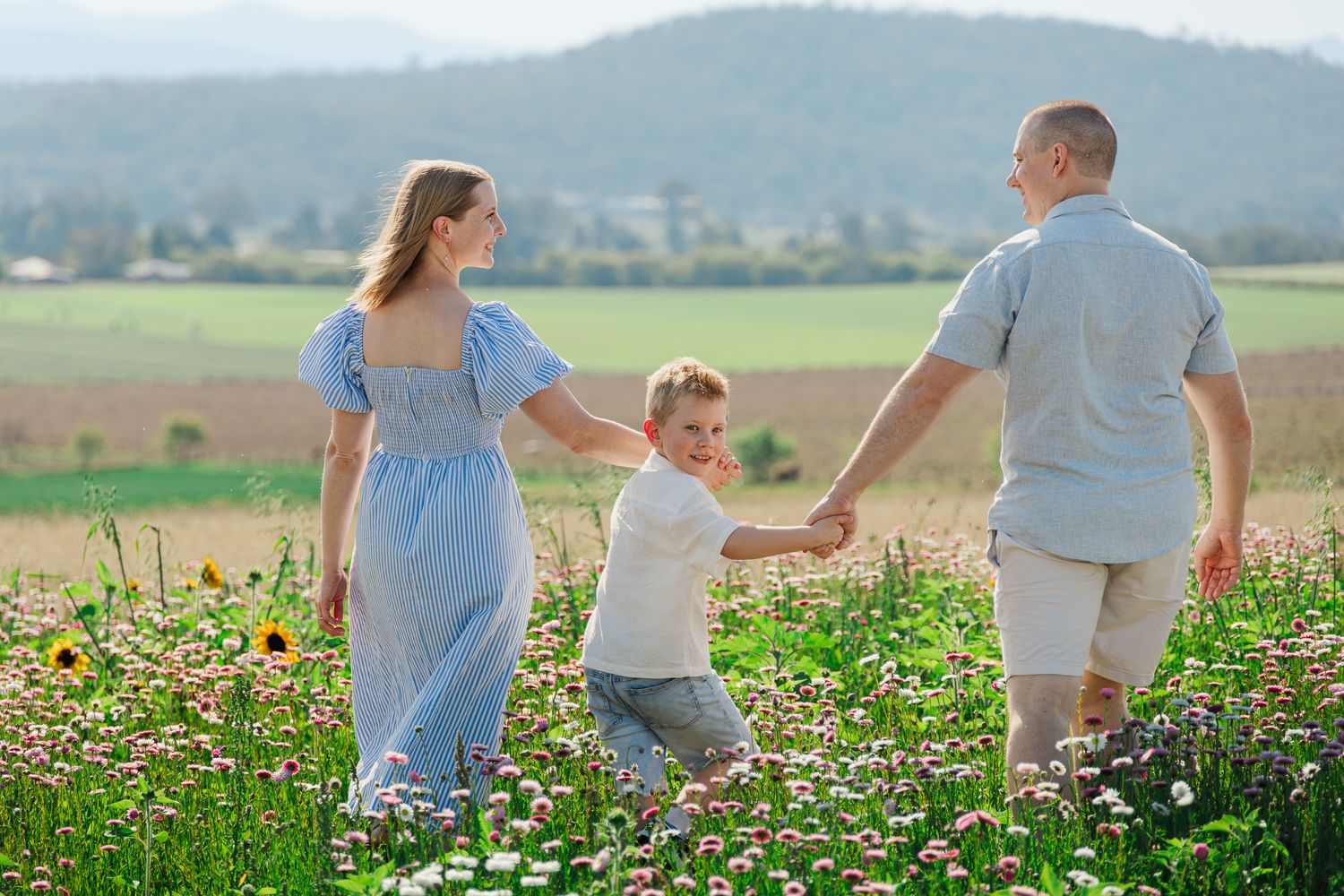Family of three holding hands in a flower field with mountains in the background on a sunny day.