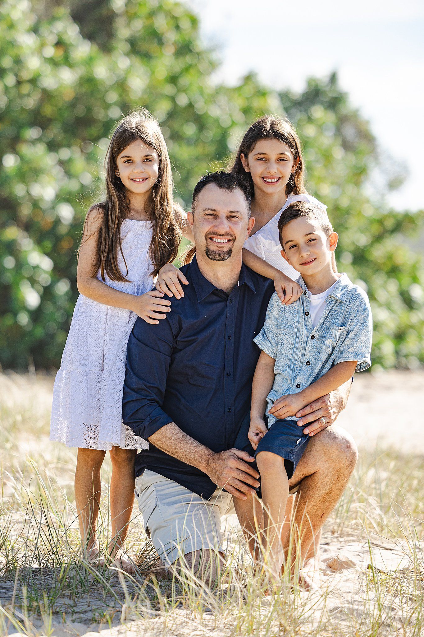A loving family poses together outdoors on a sunny day in a natural setting with green trees in the background.