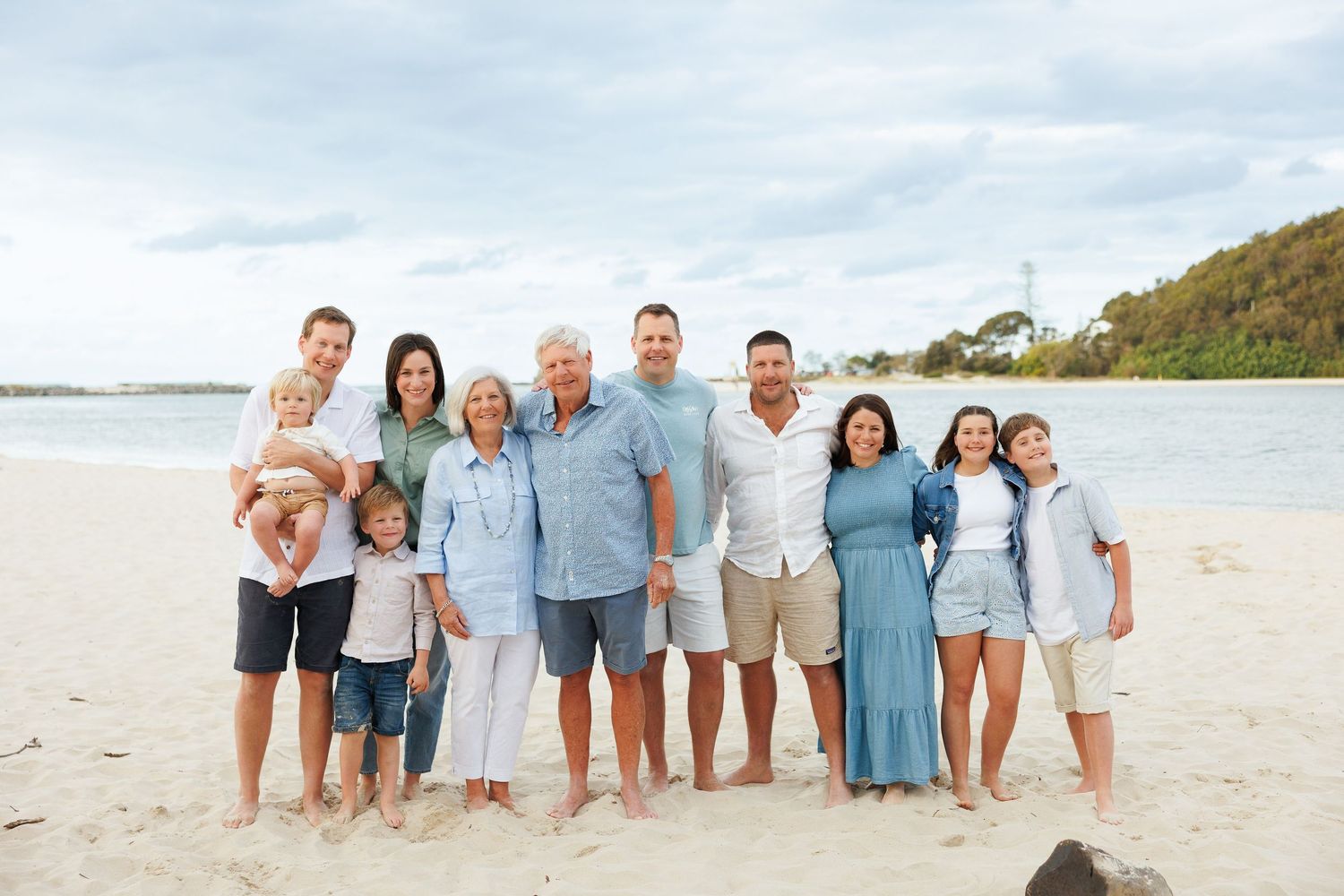 A group of people in coordinated blue and white outfits stand together on a sunny beach with water and hills in background.