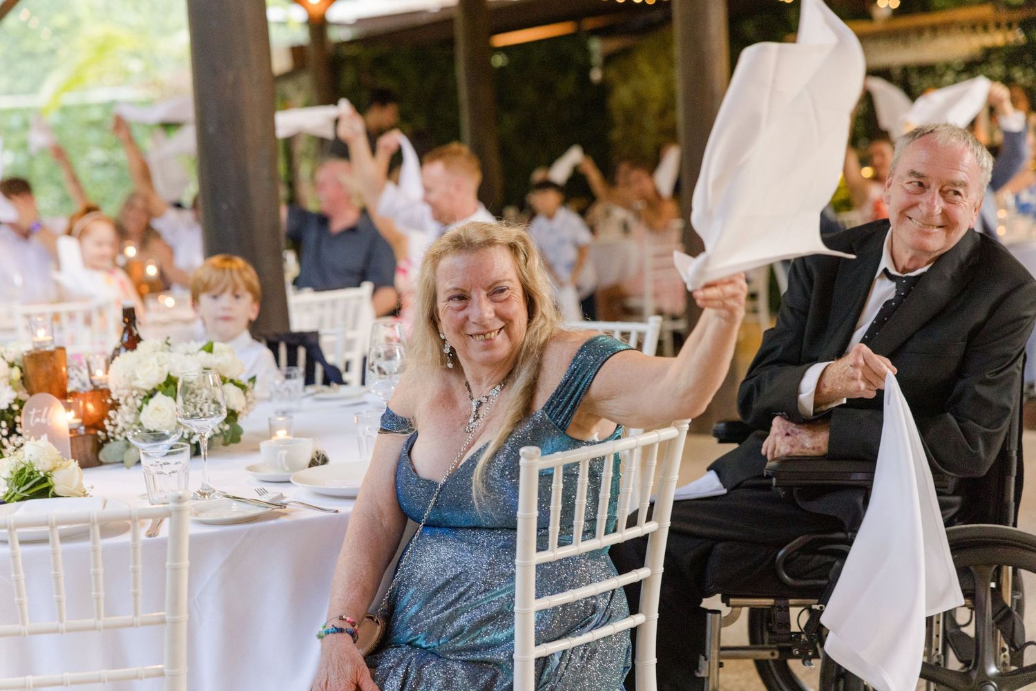 Wedding guests celebrate joyfully at an outdoor reception with white chairs and floral centerpieces.