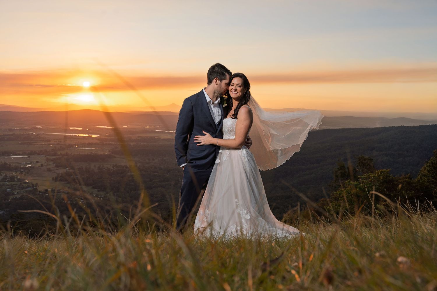 A couple embraces on a hilltop during a dramatic sunset with a flowing wedding dress veil.