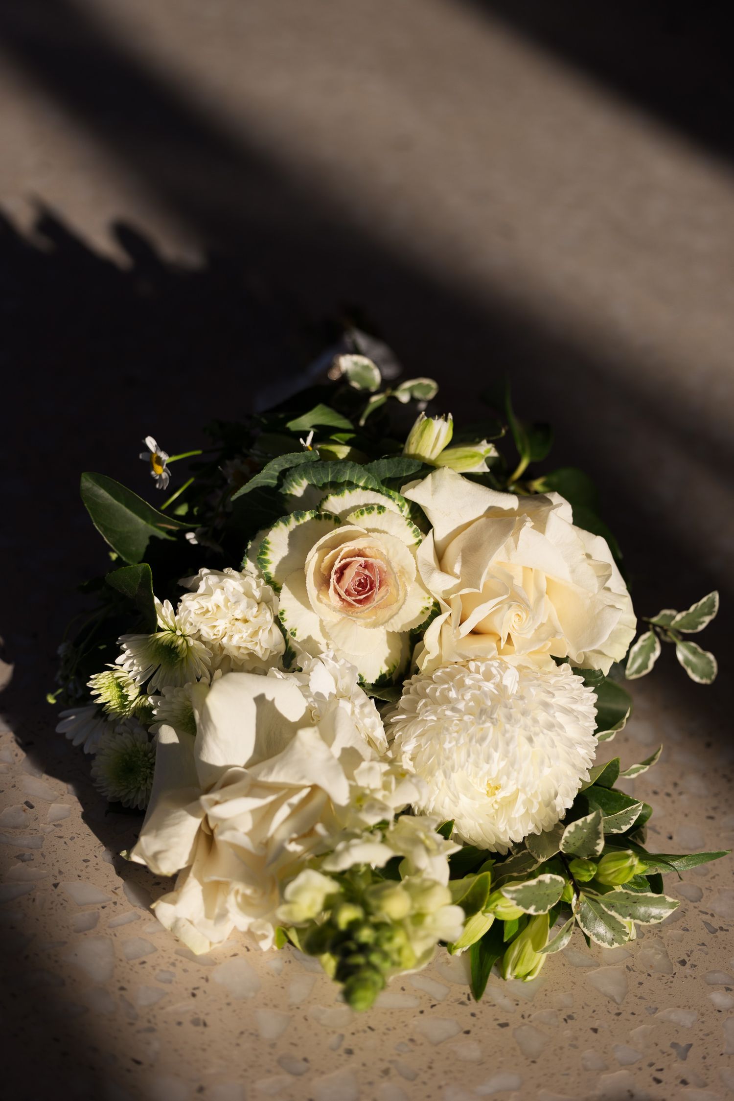 Elegant bridal bouquet of white roses, dahlias, and greenery resting on a stone surface in soft natural light.
