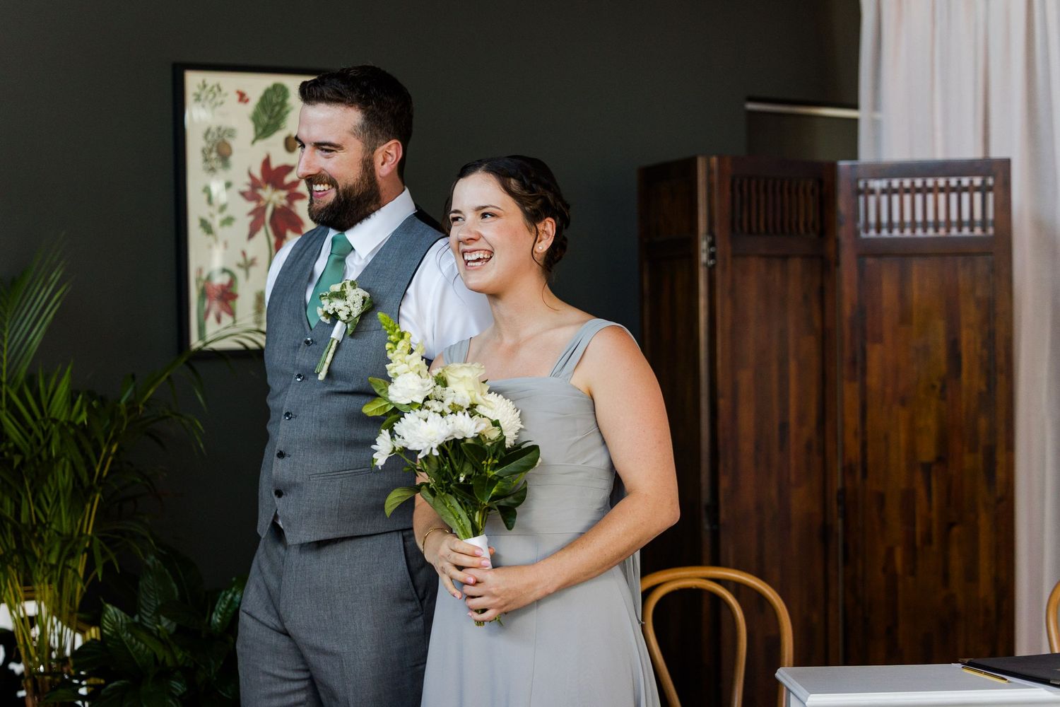 A smiling bride holding white flowers stands beside a bearded groom in a gray suit at a wedding ceremony.