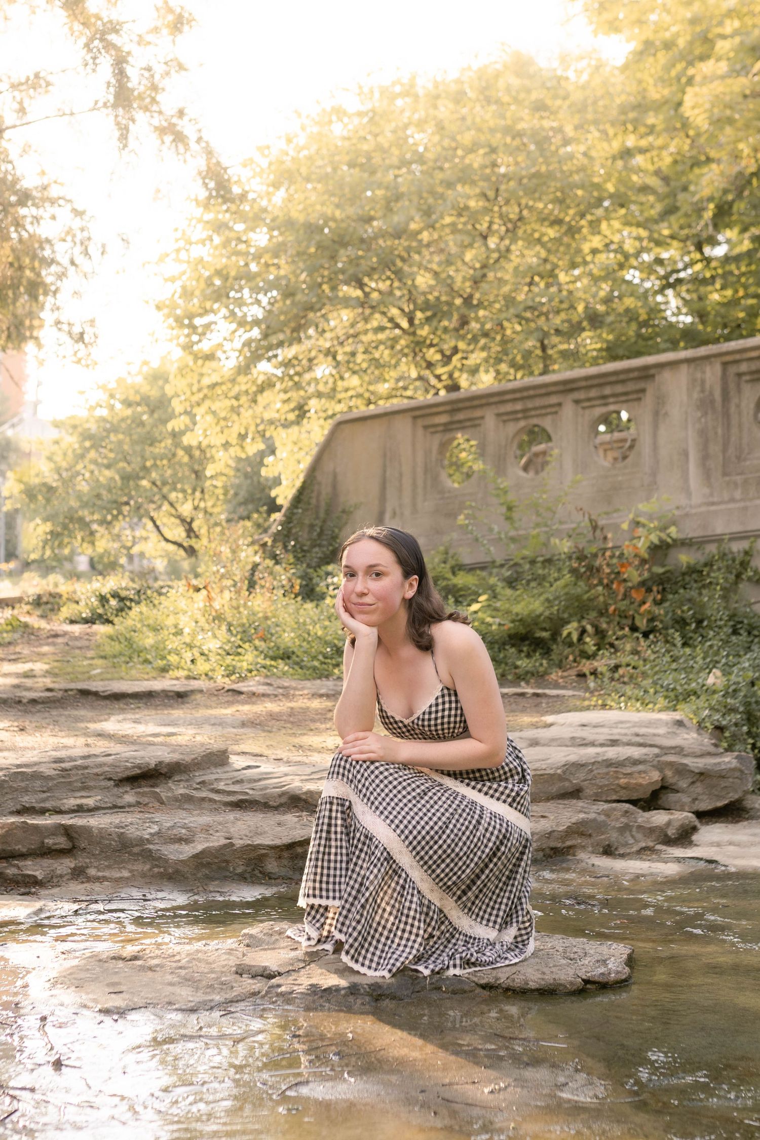 Senior girl in dress kneeling on stone in river in front of bridge