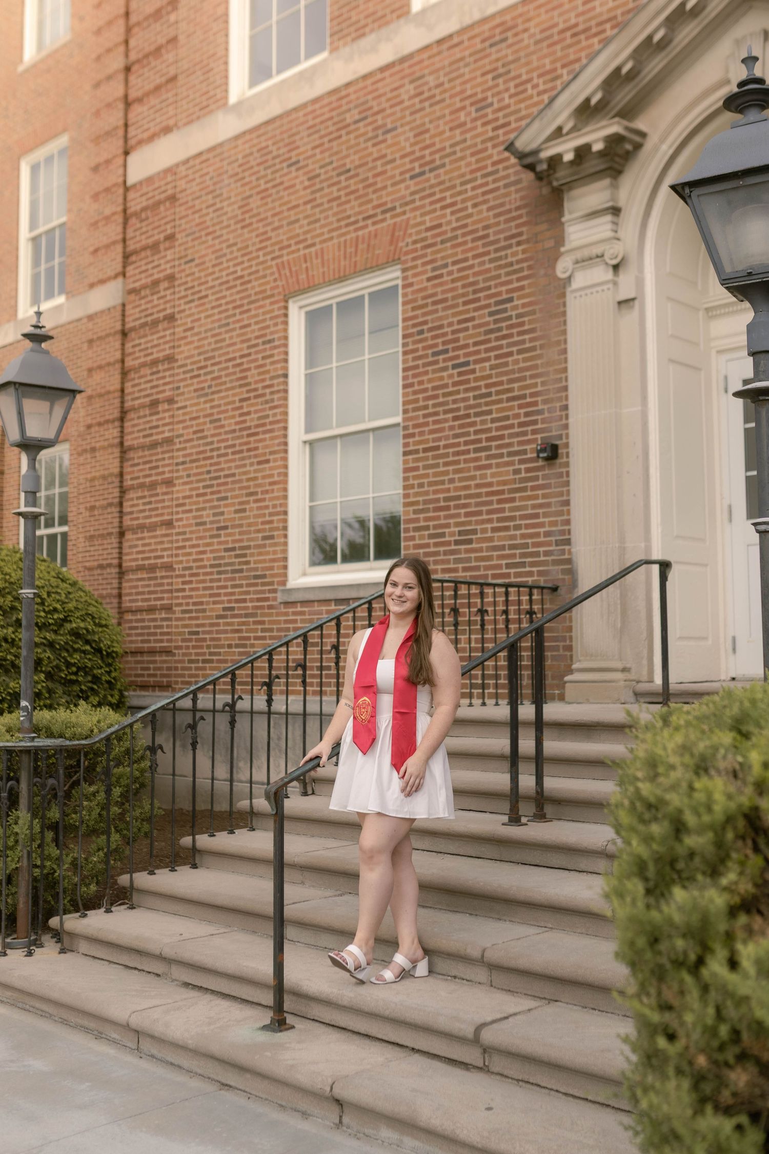 College senior standing in regalia and white dress in front of academic building