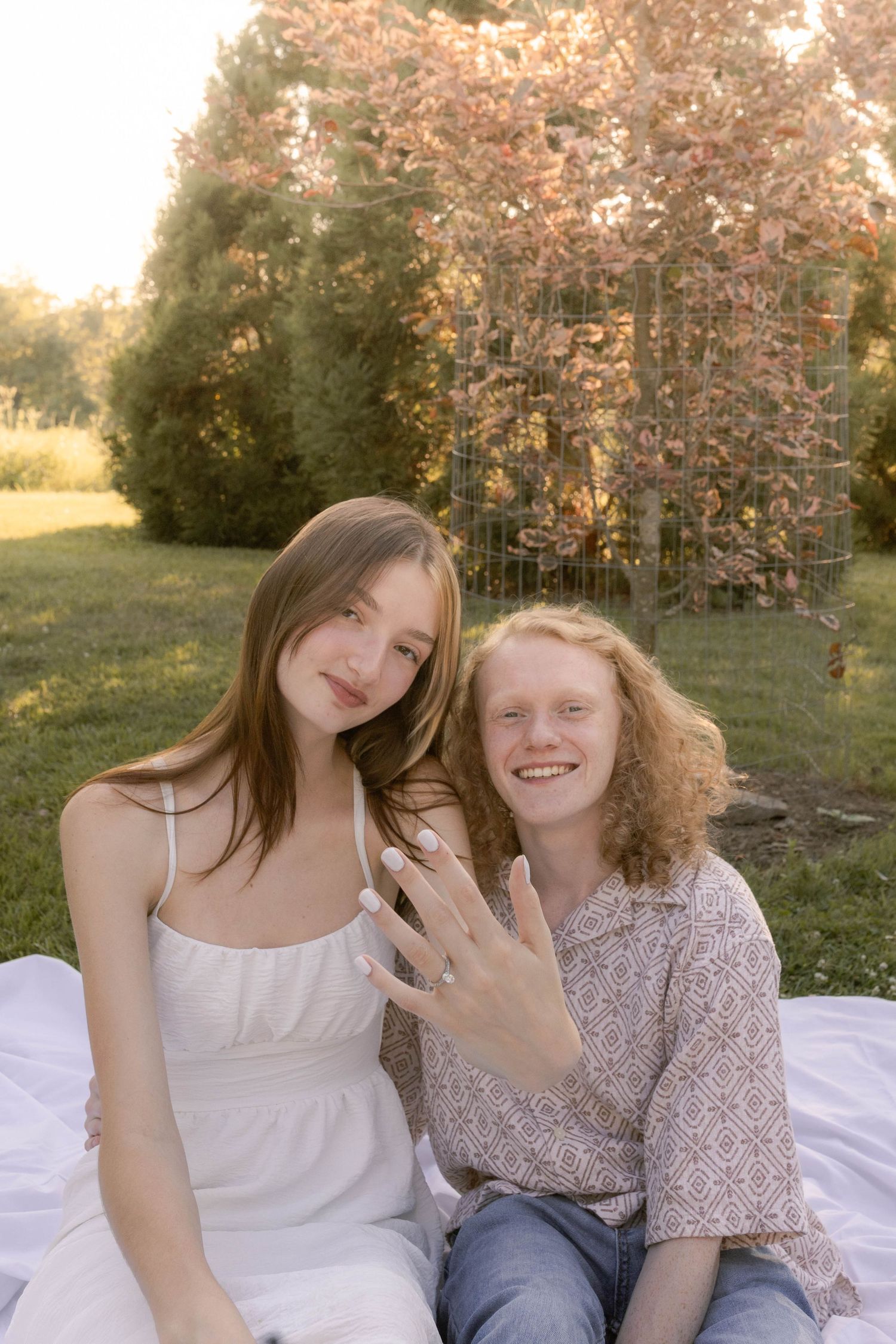Couple sitting together between trees showing off ring during sunset engagement