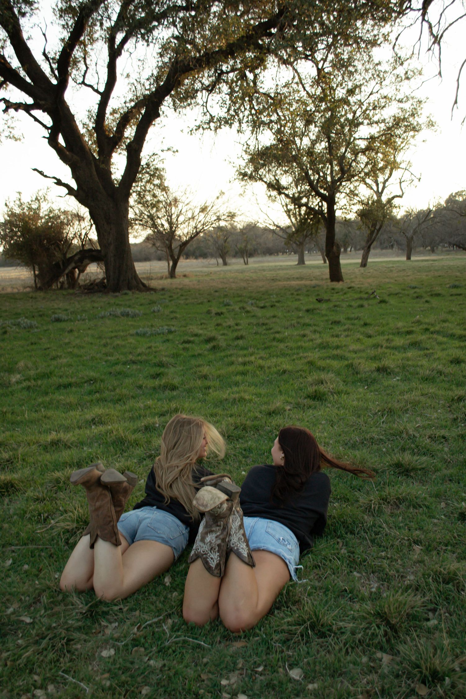 Two young women in denim shorts and cowboy boots lying on green grass chatting at sunset in a tree-lined field.
