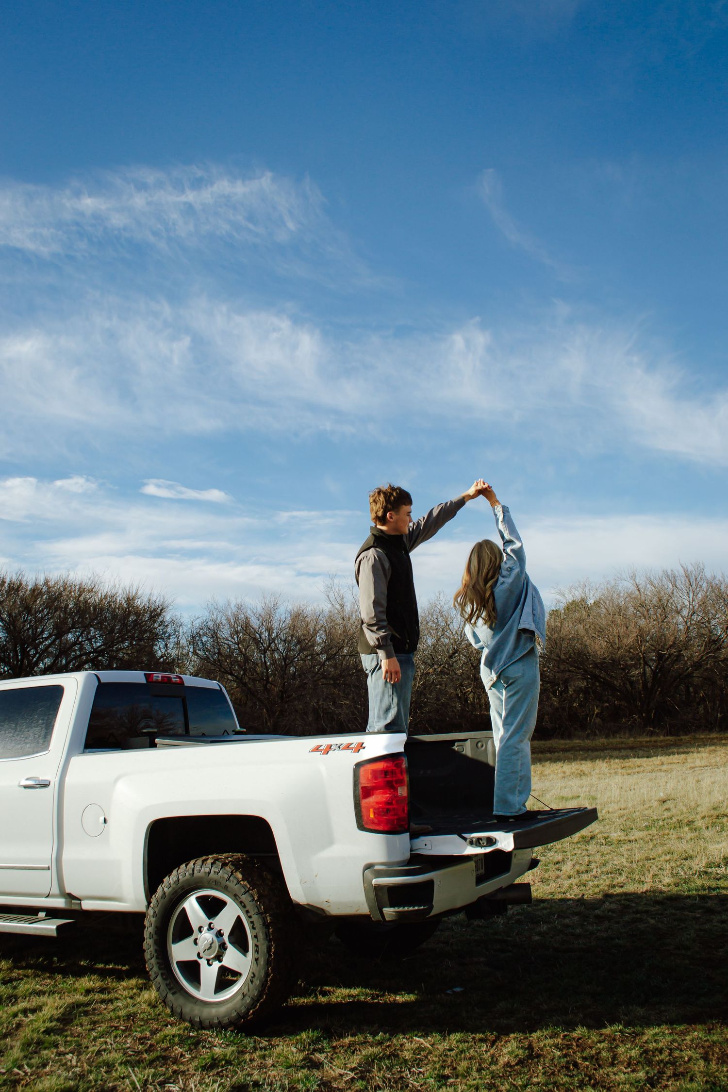 Couple dancing together standing in the bed of a white Chevy 4x4 pickup truck in a rural field under blue sky.