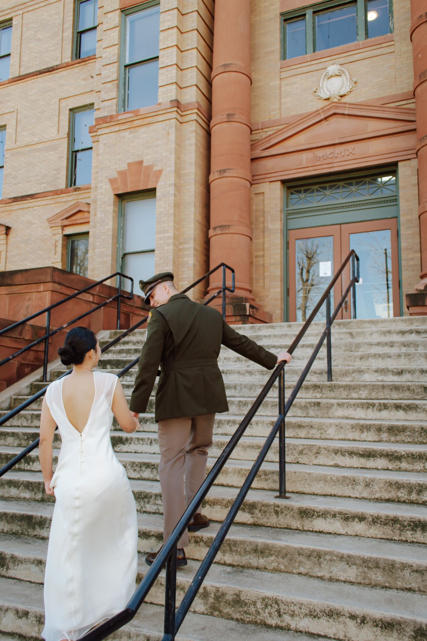 Bride in backless white gown and military groom climb stone steps of a grand historic courthouse.