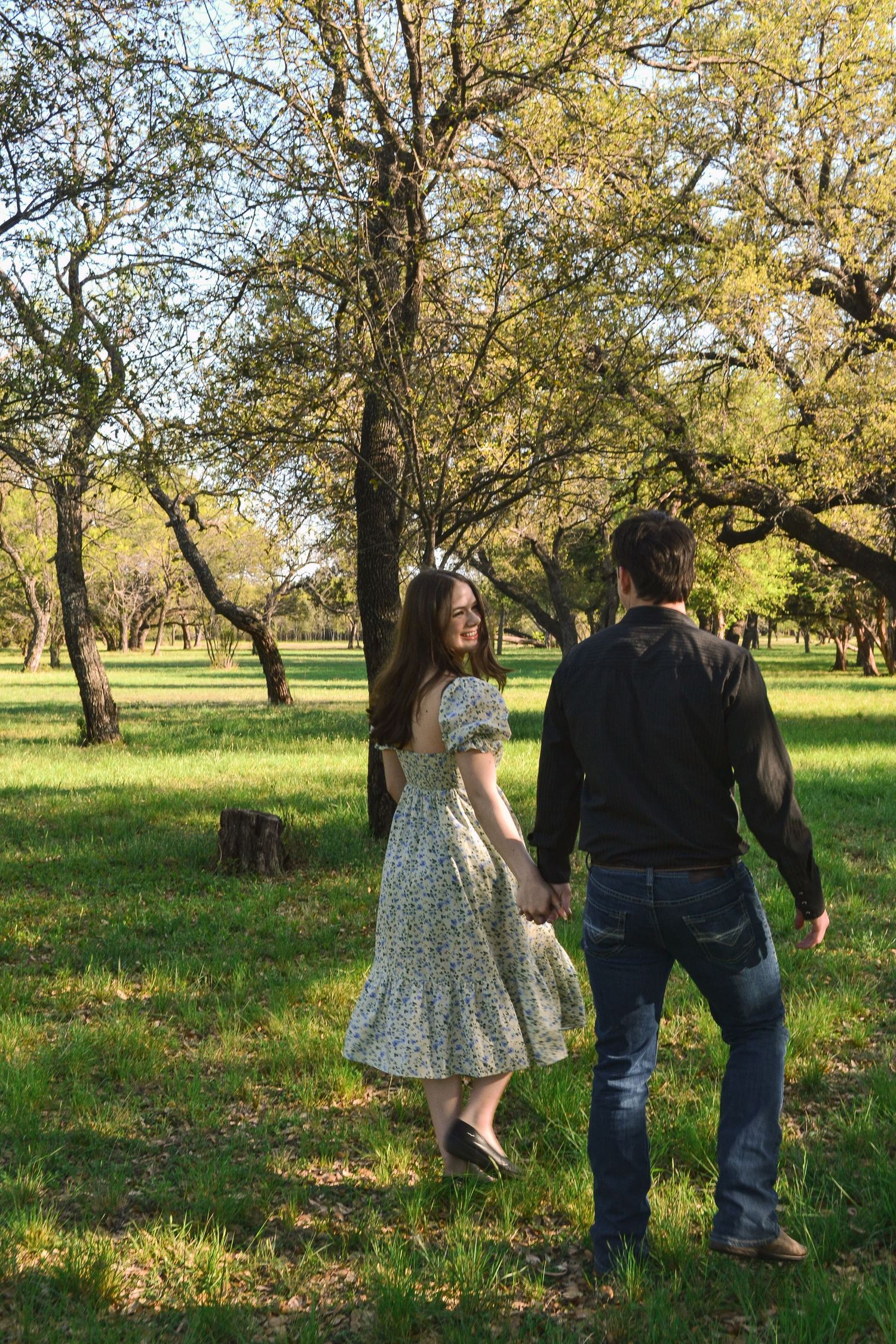 Couple holding hands walking through sunlit green park with tall trees in spring season.