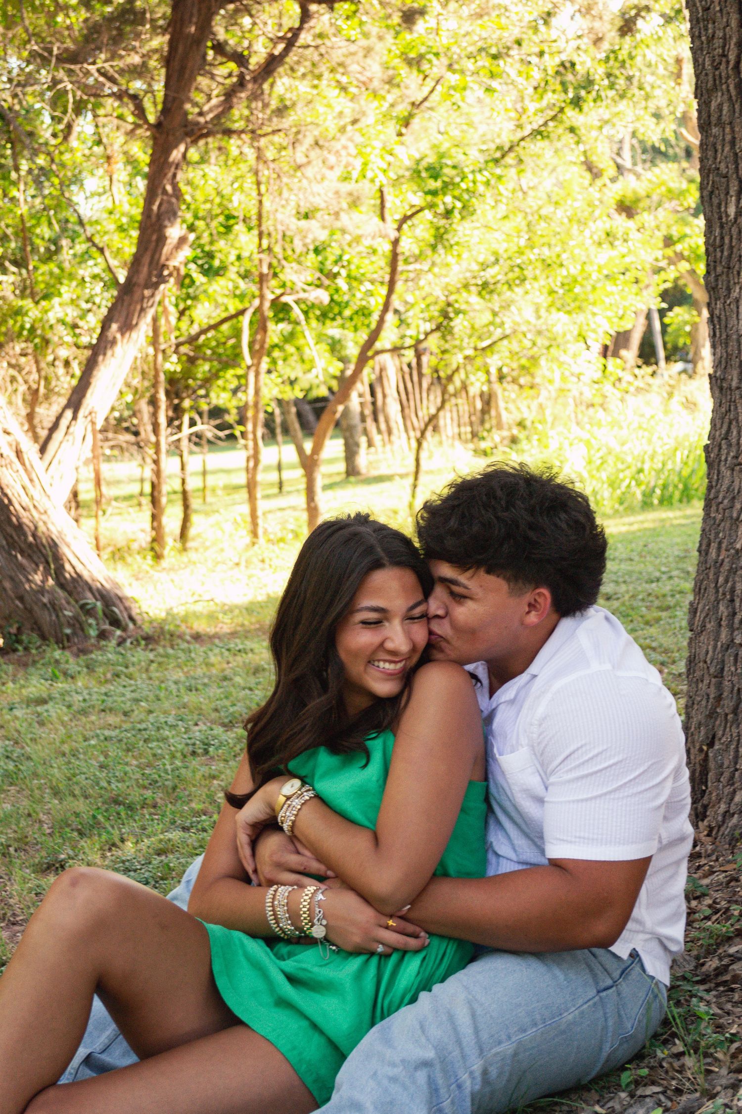 Couple sitting under a tree in a park, man kissing woman on cheek, woman in green dress smiling.