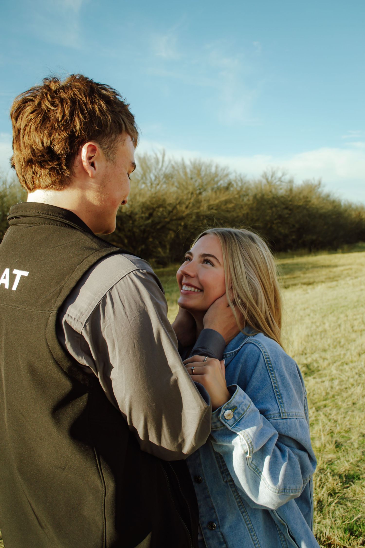 Happy young blonde woman smiling up at her boyfriend while embracing outdoors in a sunny green field.