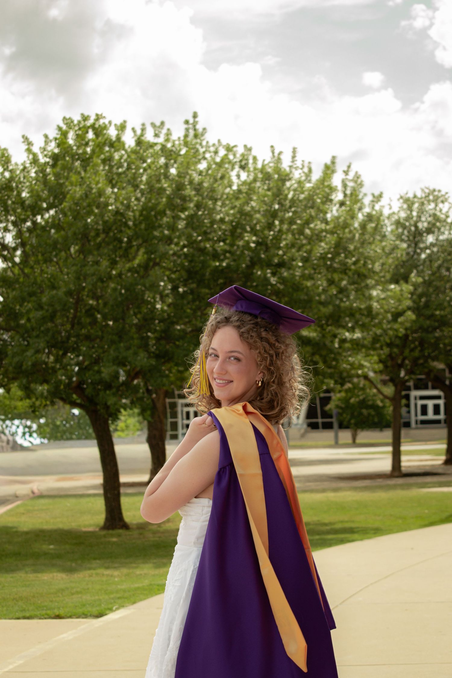 Young woman in purple graduation cap and gown smiles over her shoulder outdoors on a sunny campus walkway.