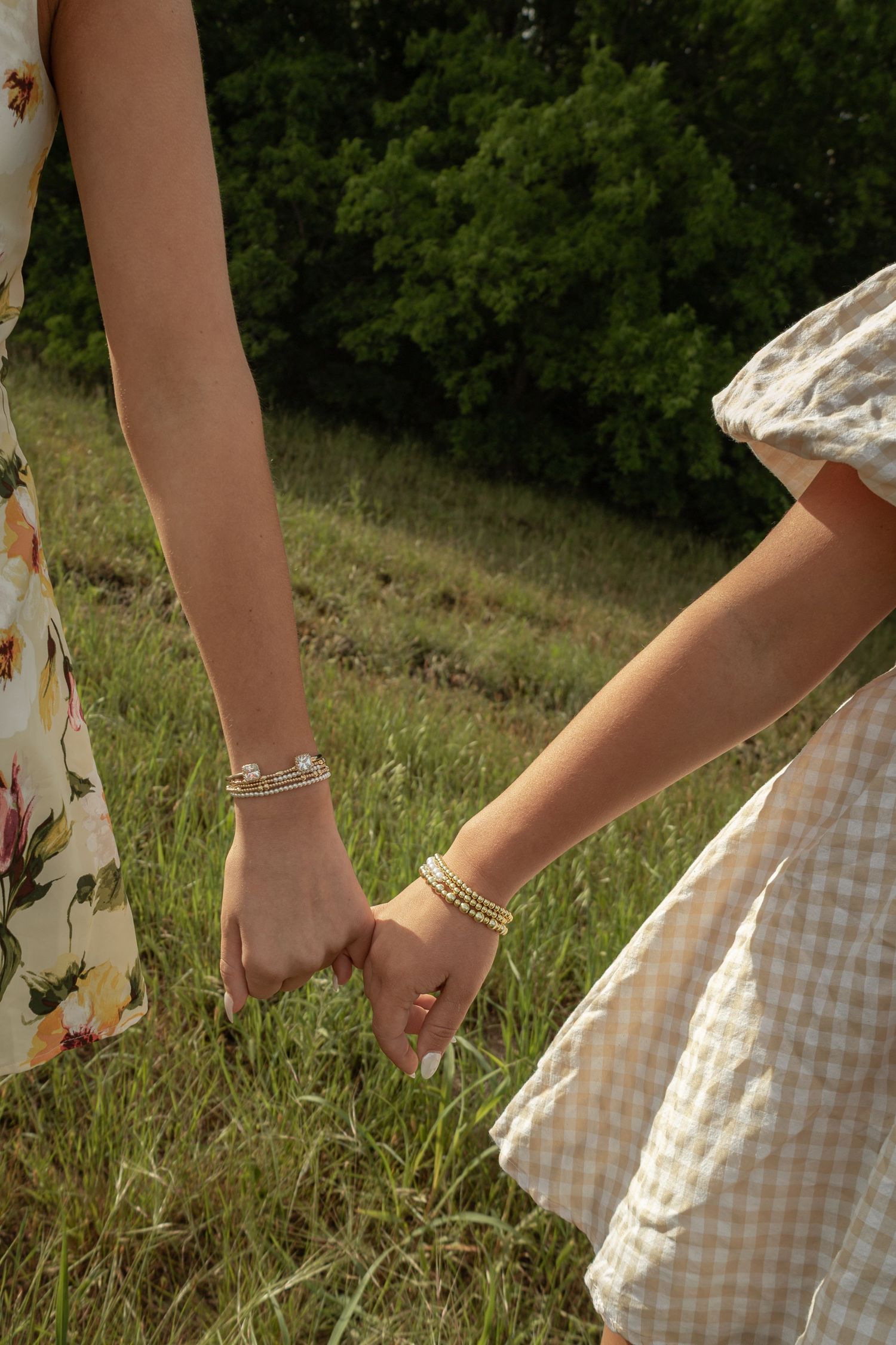 Two women holding hands in a field, wearing floral and gingham dresses with gold bracelets.