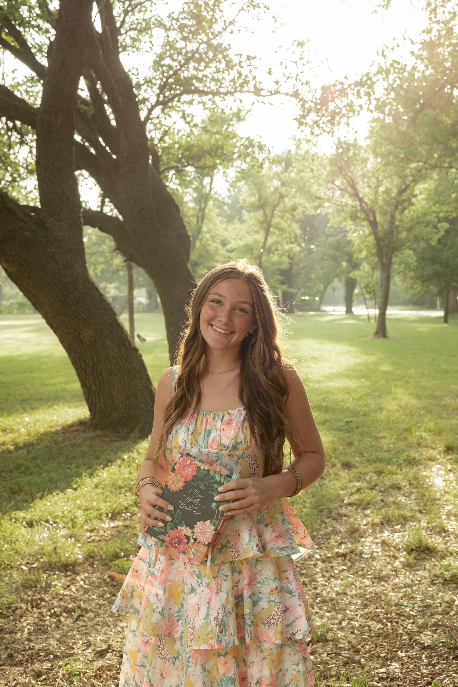 Young woman in floral dress holding a book, smiling in a sunlit park with green trees.