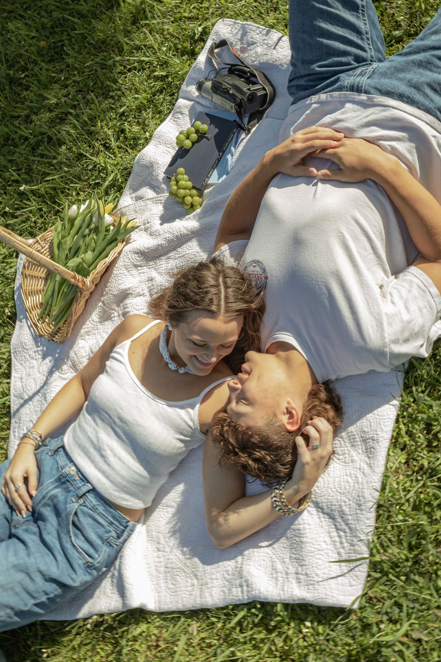Couple lying on a white picnic blanket in green grass, smiling at each other with a basket of tulips nearby.