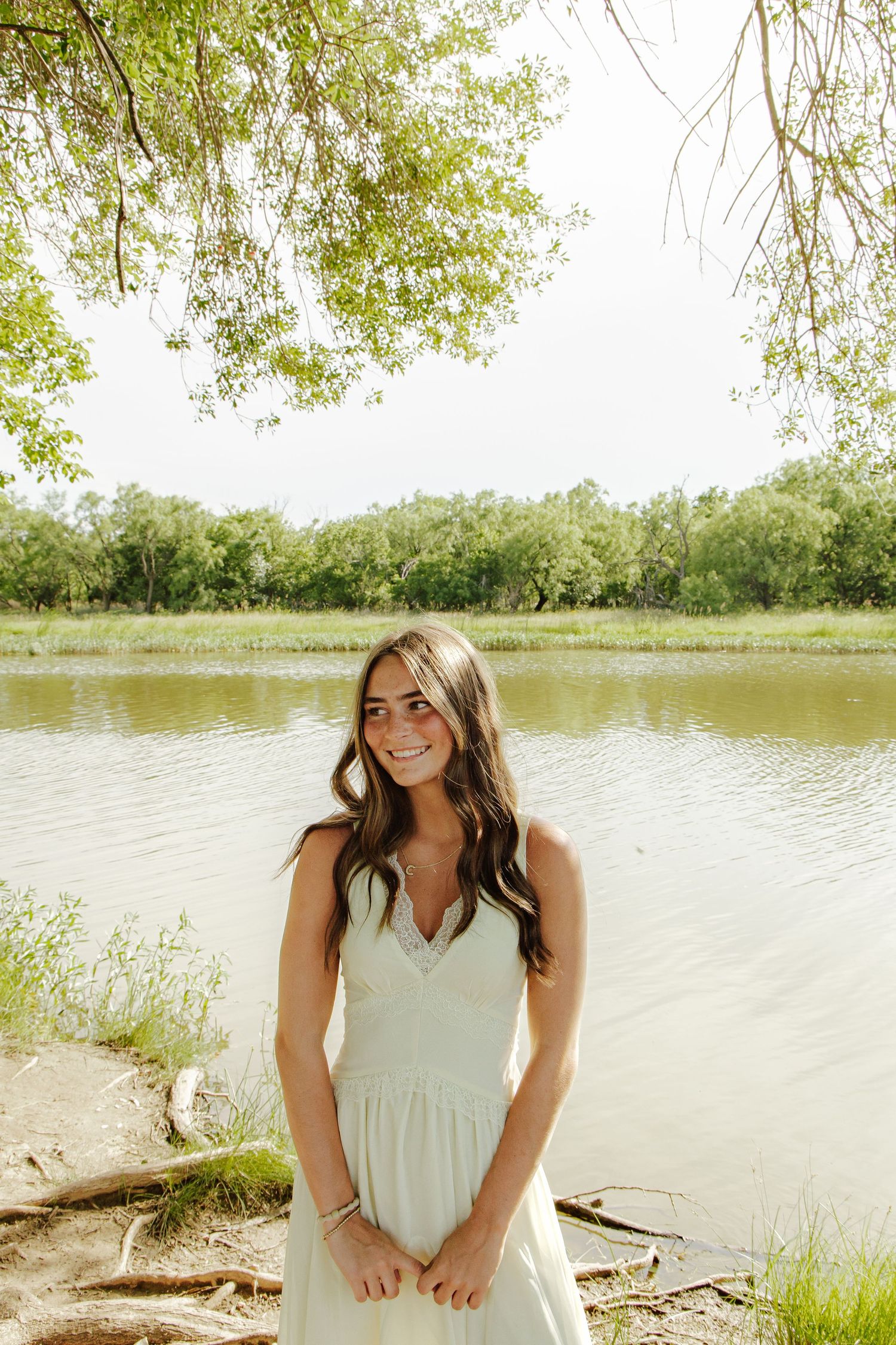 Young woman in white sundress smiling by a serene river surrounded by lush green trees on a bright summer day.