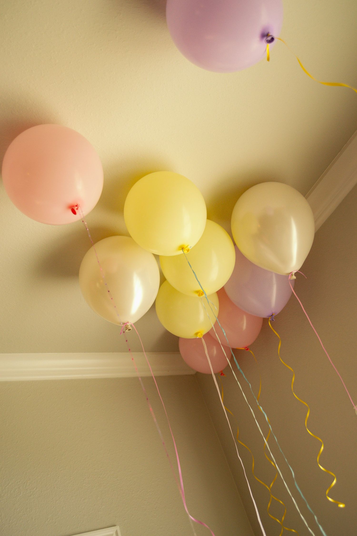 Pastel pink, yellow, and lavender helium balloons float against a ceiling corner with colorful curling ribbons.
