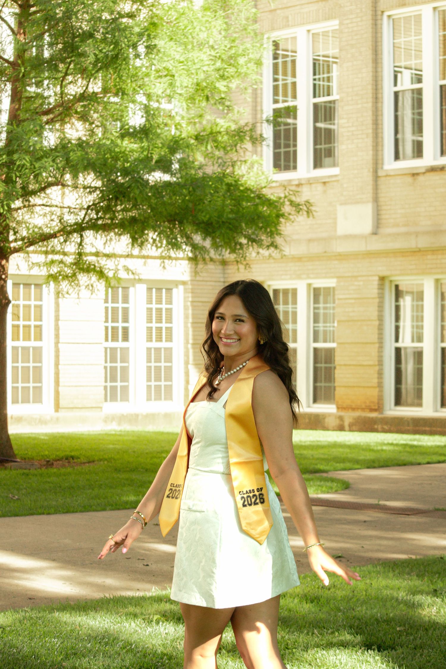 Smiling graduate in white dress and gold Class of 2023 stole poses outdoors near brick building on sunny day.