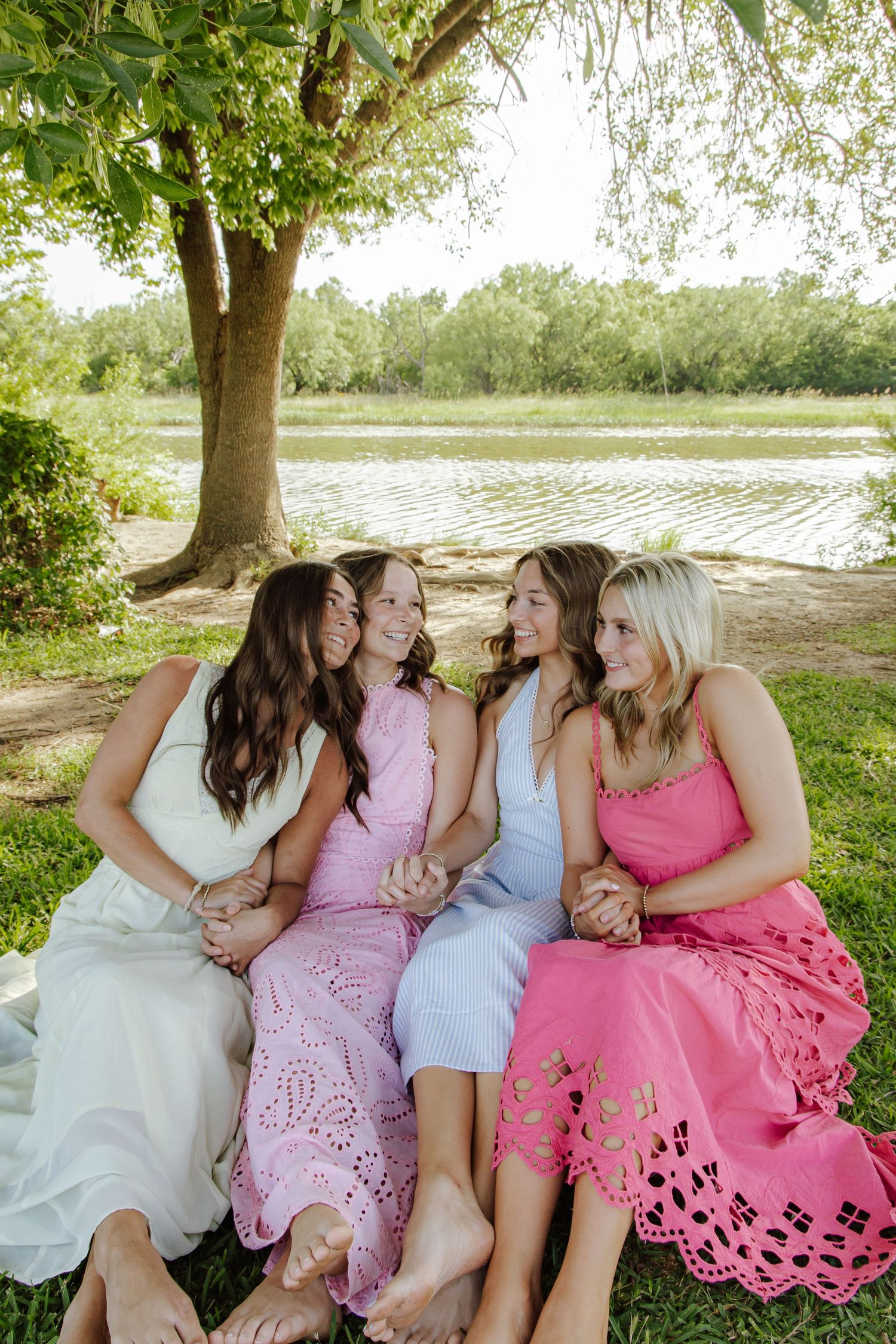 Four young women in colorful summer dresses sitting together laughing by a peaceful lakeside under a tree.