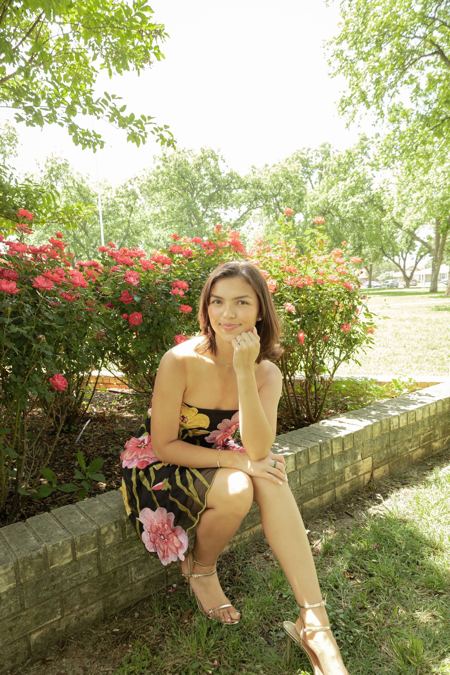 Young woman in floral dress sitting on brick ledge in park with blooming rose bushes in background.