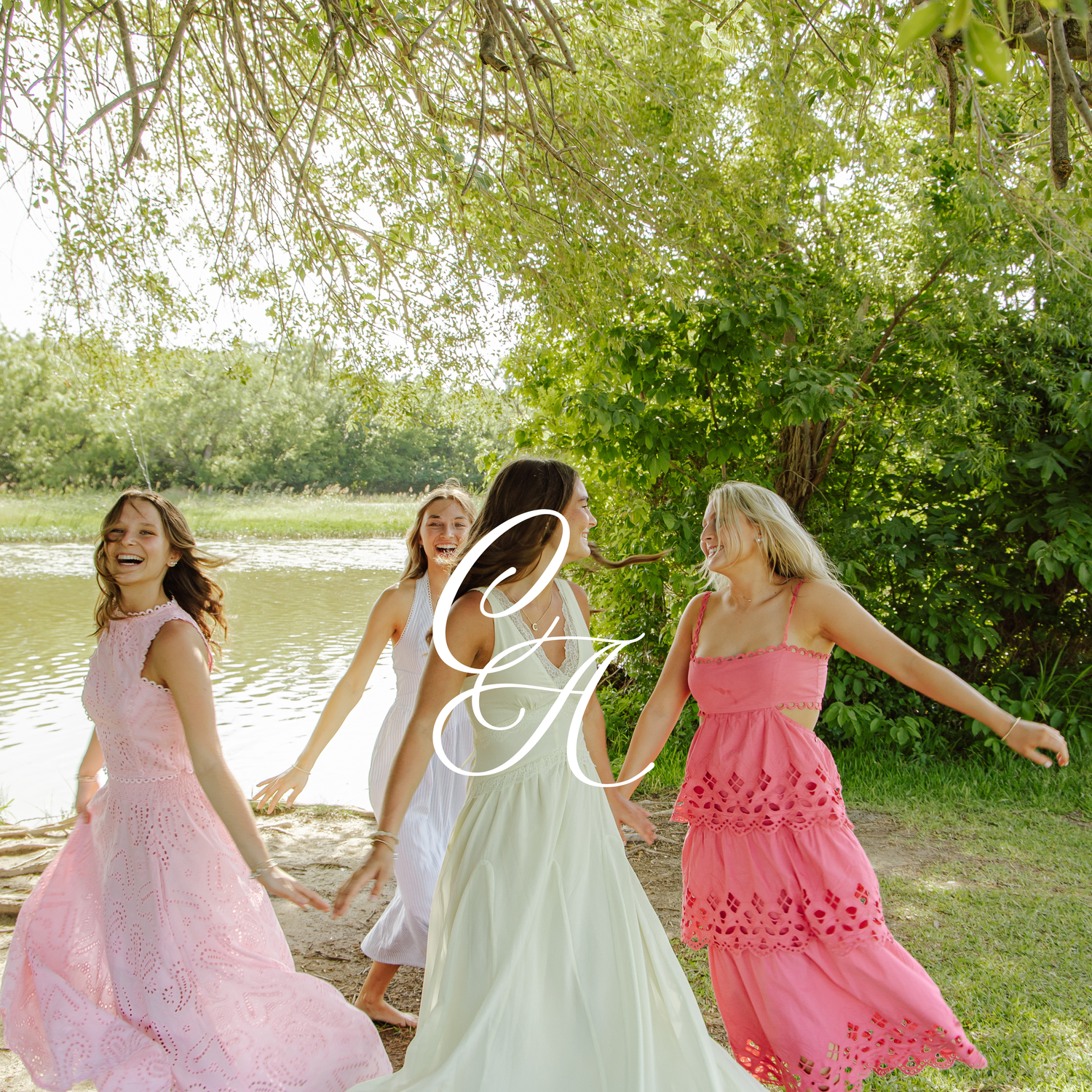 Three women in pink, white, and coral maxi dresses dancing outdoors near a serene lake surrounded by lush green trees.