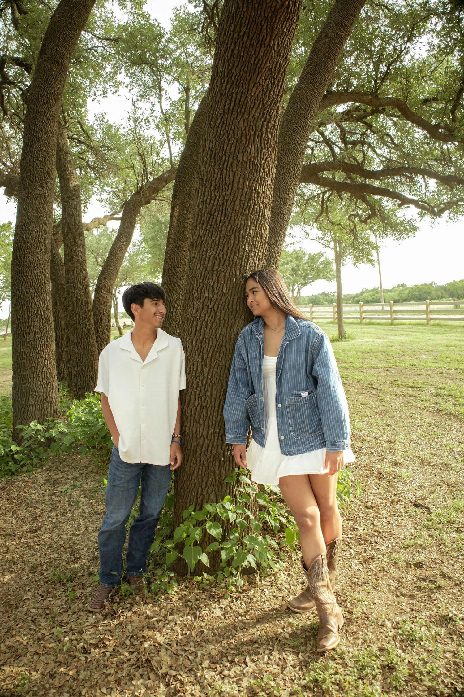 A young couple leans against a large oak tree in a park, smiling at each other in casual summer outfits.