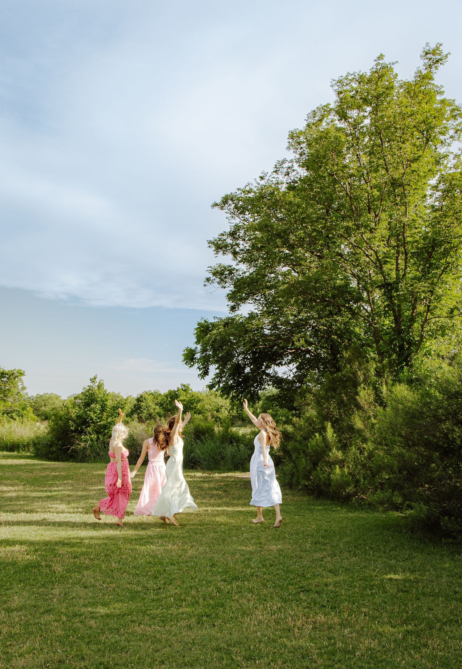 Three women in flowing dresses dance joyfully on a lush green lawn surrounded by trees under a soft blue sky.