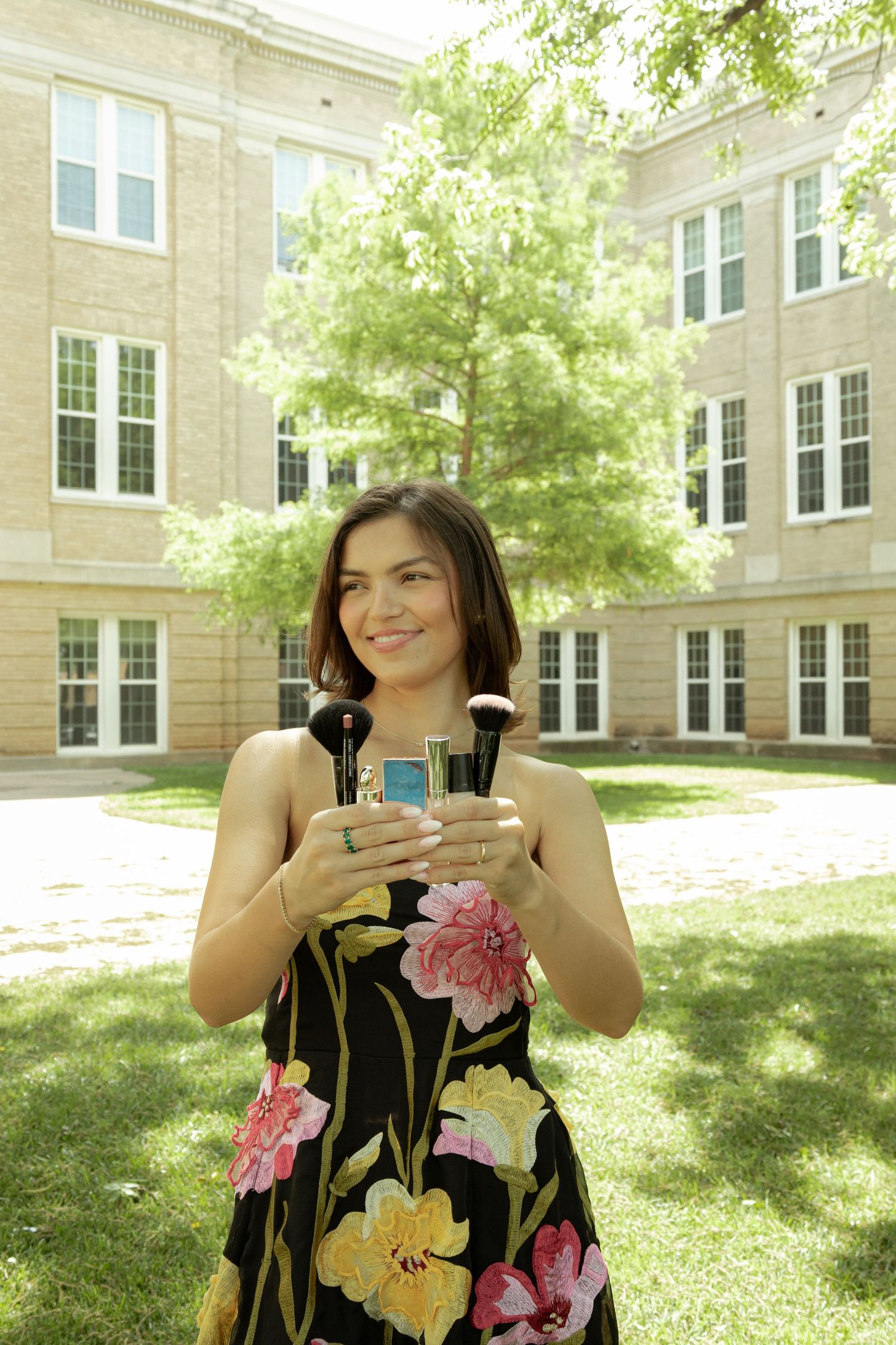 Young woman in floral dress holding makeup brushes smiling outdoors near a brick building on a sunny day.